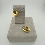 Chunky goldfarbene Tropfen-Creolen auf leinenbezogenem Podest vor hellem Hintergrund