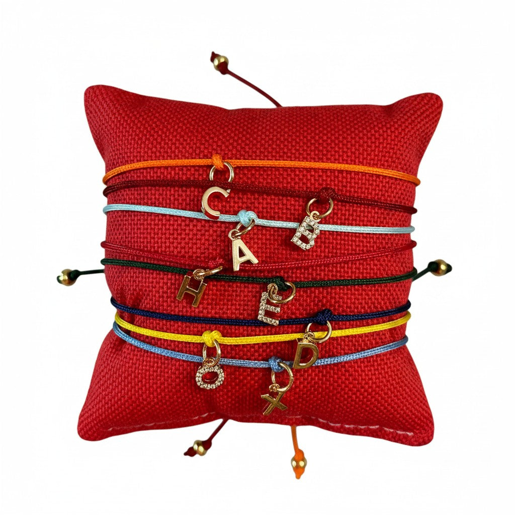 ANNA VON BERGMANN Armbänder mit goldenen Buchstabenanhängern und Zirkonia an bunten Nylonkordeln auf rotem Kissen