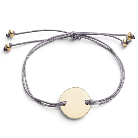 ANNA VON BERGMANN CORD Armband mit goldplattierter runder Plakette an einem grauen Nylonband auf weißem Hintergrund